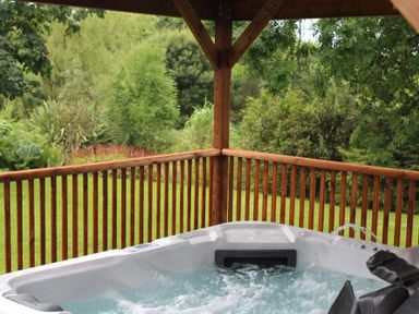 An outdoor hot tub on a wooden deck surrounded by green trees and plants at Pond Cottage in New Quay Ceredigion
