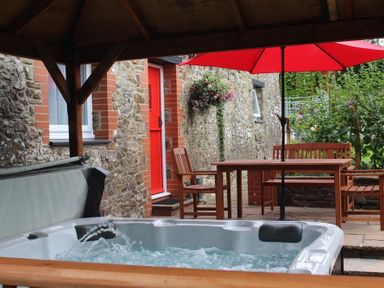 An outdoor patio area with a hot tub a wooden table and chairs with a red umbrella attached to the table next to a stone wall with a red door and window at Pond Cottage in New Quay Ceredigion