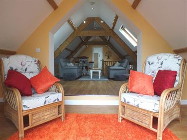 A living room with wicker chairs, red and orange cushions, wooden beams, and grey sofas at Pond Cottage in New Quay Ceredigion