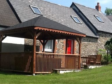 A stone cottage with a red door and a wooden gazebo on a grass lawn at Pond Cottage in New Quay Ceredigion