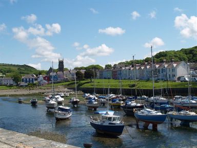 A harbor with boats docked in water near a row of houses and a church in the background at Dewi Villa in Aberarth Ceredigion