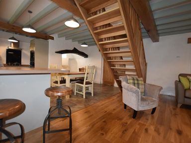 A dining area with wooden chairs and tables next to a kitchen counter with stools and a wooden staircase at The Old Farmhouse Kitchen in St Davids Pembrokeshire