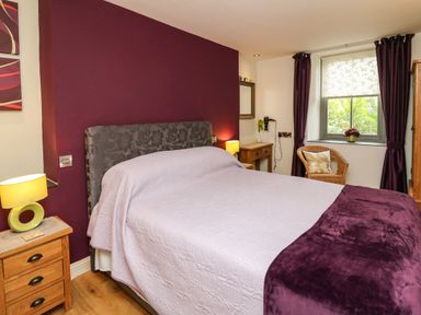 A bedroom with a bed bedside tables two lamps a wicker chair a window with curtains and a desk at Nevern House in Newport Pembrokeshire