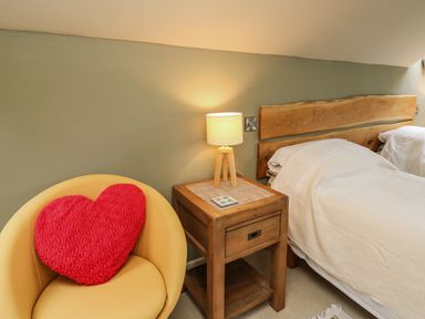 A bedroom with a wooden bedside table and lamp next to a bed with a wooden headboard and a yellow chair with a red heart pillow at Nevern House in Newport Pembrokeshire