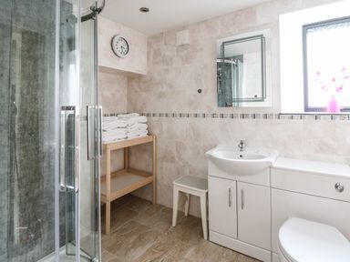 A bathroom with a glass shower enclosure white sink and toilet wooden shelf with towels and a wall clock at Nevern House in Newport Pembrokeshire