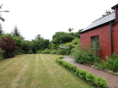 A garden with a mowed lawn a paved path shrubs a red building and trees at Nevern House in Newport Pembrokeshire