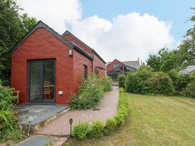A red building with large glass doors and a path leading to another house with a lawn and bushes at Nevern House in Newport Pembrokeshire