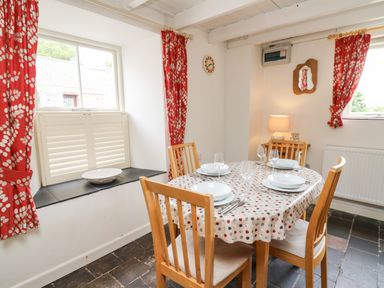 A dining room with a table set with plates and glasses red curtains on windows and wooden chairs at Panteg Cottage Newport