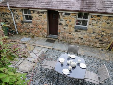 An outdoor stone patio with a table set for tea and four chairs in front of a stone building with a wooden door and two windows at Panteg Cottage Newport Pembrokeshire