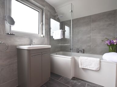 A bathroom with a bathtub and shower, a sink cabinet, a round mirror, a window, towels, and a vase of flowers at Greenfields in St Davids Pembrokeshire