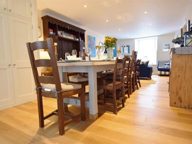 A dining room with a long table and wooden chairs a dark wooden cabinet and a living area in the background at Gwelfor Newport in Newport Pembrokeshire