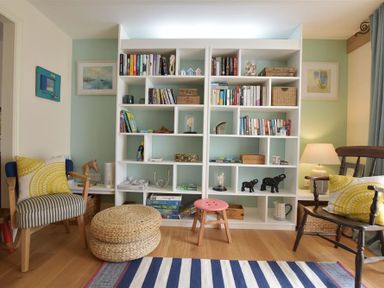 A room with a white bookshelf filled with books and decorative items two chairs with yellow patterned cushions a striped rug and a table lamp at Gwelfor Newport