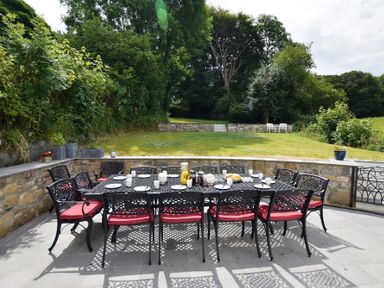 An outdoor dining area with a metal table and eight chairs on a patio overlooking a lawn and trees at Gwelfor Newport