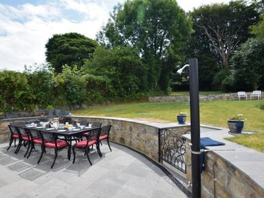 An outdoor patio with a dining table and chairs with red cushions and a grassy garden with trees and stone wall at Gwelfor Newport in Newport Pembrokeshire