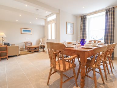 A dining area with a wooden table set with white dishes and glasses next to a window with curtains and an adjacent living room with sofas and wicker furniture at Neverndale in Newport Pembrokeshire