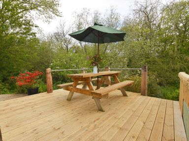 A wooden picnic table with a green umbrella and flowers on a wooden deck surrounded by trees at Mill Barn Denant in Haverfordwest Pembrokeshire