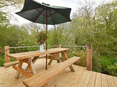 A wooden picnic table with benches and a green umbrella on a deck surrounded by trees at Mill Barn Denant in Haverfordwest Pembrokeshire