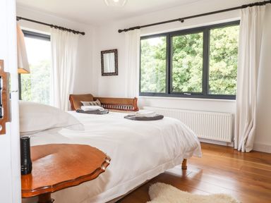 A bedroom with a wooden side table bed with white bedding two towels on bed and large windows with white curtains at Ty Cerrig in Newport Pembrokeshire