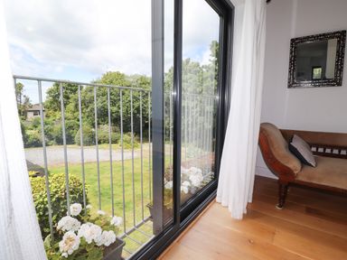 A room with wooden floor and a brown upholstered chaise lounge next to a large sliding glass door with white curtains and flowers outside the door at Ty Cerrig Newport Pembrokeshire