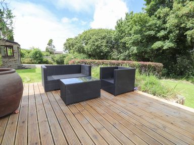 An outdoor wooden deck with black wicker furniture including a sofa armchair and table surrounded by grass and trees at Ty Cerrig in Newport Pembrokeshire
