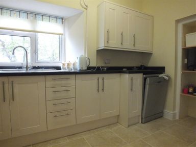A kitchen with cream cabinets black countertop a sink under a window a kettle canisters and a dishwasher with shelving in the corner at Tremynydd Fawr in St Davids Pembrokeshire