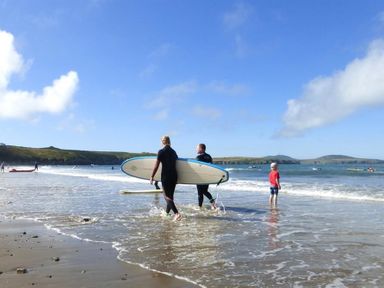 People walking in shallow water on a beach with surfboards and a child standing at the shoreline at Tremynydd Fawr in St Davids Pembrokeshire