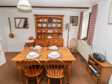 A dining room with a wooden table set for four people wooden chairs a wooden cabinet with plates and jars a floor lamp and a small shelf with a radio at Ty Canol in Mathry Pembrokeshire
