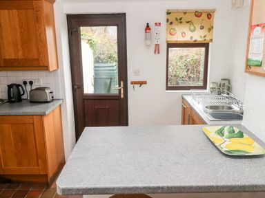 A kitchen with wooden cabinets a countertop with a tray a door and a window with fruit patterned blind at Ty Canol in Mathry Pembrokeshire