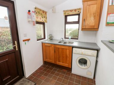 A utility room with a sink, washing machine, wooden cabinets, windows with fruit patterned blinds and a door at Ty Canol Mathry Pembrokeshire