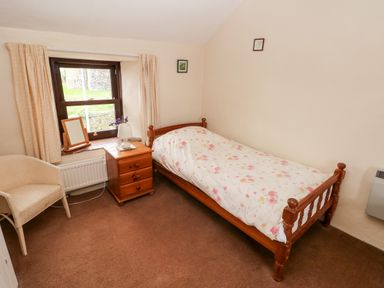 A small bedroom with a single bed with floral bedding a wooden nightstand a table lamp a window with curtains and a chair at Ty Canol Mathry Pembrokeshire