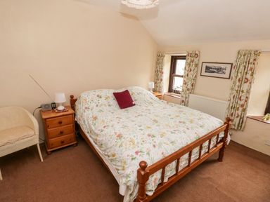 A bedroom with a wooden bed covered in floral bedding bedside tables with lamps windows with floral curtains and a wicker chair at Ty Canol in Mathry Pembrokeshire