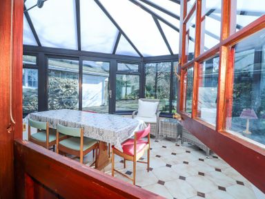 A conservatory with a table covered with a patterned cloth surrounded by chairs and a wicker chair with cushions at Meadowlands in Dinas Cross Pembrokeshire