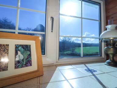 A window with a view of green fields and trees with a framed picture and a lantern on the tiled windowsill at Meadowlands in Dinas Cross Pembrokeshire