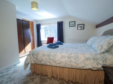 A bedroom with a bed covered with patterned bedding and towels on top a wardrobe and a window with dark curtains with a chair beneath at Meadowlands in Dinas Cross Pembrokeshire