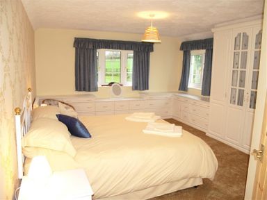 A bedroom with a bed with cream bedding and blue pillows a bedside table with a lamp wardrobes and drawers beneath two windows with blue curtains at Sarnau in Efailwen Carmarthenshire