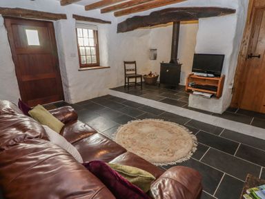 A living room with a leather sofa and cushions a wood burning stove a small round rug a wooden chair and a television on a wall shelf at Panteurig Cottage Strumble Head Goodwick Pembrokeshire