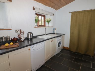 A kitchen with white cabinets black countertops a sink a washing machine a dishwasher a kettle two vases with flowers and a green curtain at Panteurig Cottage in Strumble Head Goodwick Pembrokeshire