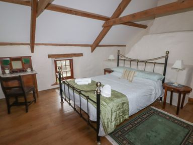 A bedroom with a metal bed frame green bedspread wooden beams wooden floor a small window a wooden desk with chair and two bedside tables with lamps at Panteurig Cottage Strumble Head Goodwick Pembrokeshire
