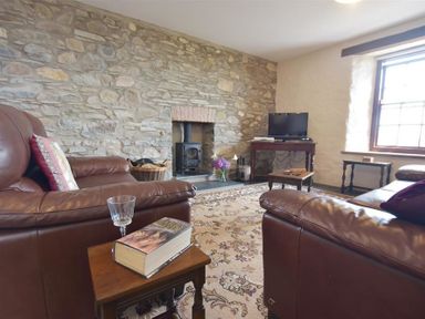 A living room with brown leather sofas a stone wall with a fireplace a wooden TV stand with a television and a window at Panteurig Farmhouse in Strumble Head Goodwick Pembrokeshire