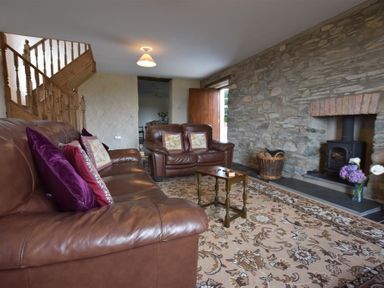 A living room with brown leather sofas a small wooden table a stone wall fireplace a basket of firewood and a floral carpet at Panteurig Farmhouse in Strumble Head Goodwick Pembrokeshire
