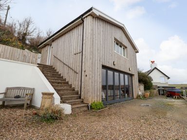 A wooden building with stairs on the side and a gravel driveway at Blackrock Boathouse in Dale Pembrokeshire