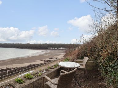 An outdoor seating area with two wicker chairs and a round glass table overlooking a sandy beach and water at Blackrock Boathouse in Dale Pembrokeshire