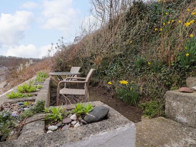 An outdoor seating area with a table and two chairs next to a hillside with plants and yellow flowers at Blackrock Boathouse in Dale Pembrokeshire