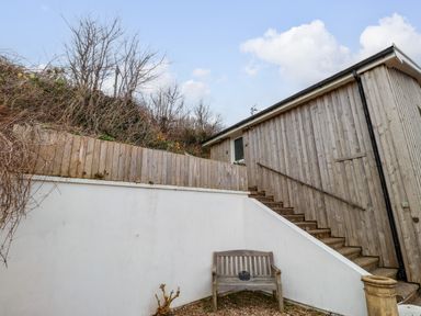 An outdoor area with wooden stairs leading to a door and a wooden bench against a white wall at Blackrock Boathouse in Dale Pembrokeshire