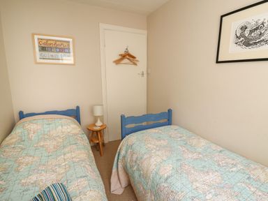 A bedroom with two single beds with maps on the bedcovers a small wooden round table with a lamp and a striped pillow on one bed at Yr Odyn in Llangrannog Ceredigion