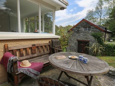 An outdoor patio with a wooden table and bench with a pink throw and hat next to a stone building at Yr Odyn in Llangrannog Ceredigion