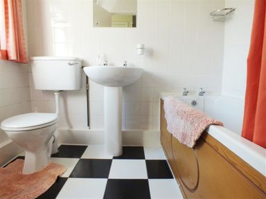 A bathroom with a toilet, pedestal sink, bathtub with wooden panel and black and white tiled floor at Granston Cottage in Castlemorris Pembrokeshire