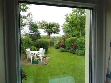 A view of a garden with a white table and chairs on a patio surrounded by bushes and trees at Granston Cottage in Castlemorris Pembrokeshire