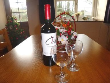 A bottle of wine and two empty wine glasses on a wooden table near a floral decoration and a window at Granston Cottage in Castlemorris Pembrokeshire