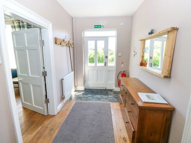 An entrance hallway with a wooden dresser and mirror on the right wall a coat rack on the left wall and a white door with a fire exit sign above at Newton West Farm Newton near Newgale